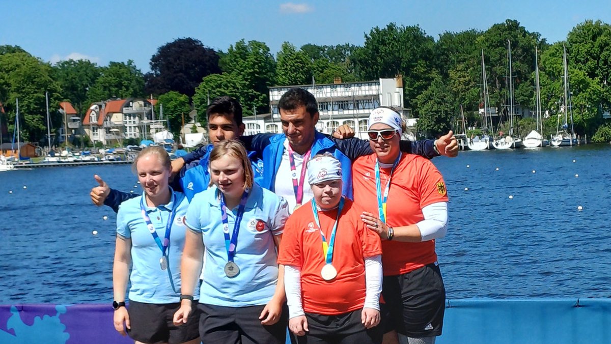 Ahtletinnen und Athleten stehen mit ihren Medaillen an einem Ufer. Im Hintergrund sind Boote zu sehen. 