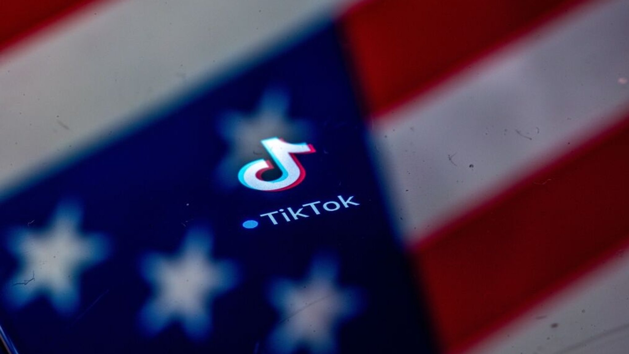 ILLUSTRATION - 10.04.2025, Mecklenburg-Vorpommern, Schwerin: Das auf einem Bildschirm angezeigte Logo der Videoplattform TikTok, einer Social-Media-App des chinesischen Technologiekonzerns Bytedance, erscheint auf einem Display, in dem sich die US-amerikanische Flagge spiegelt. (zu dpa: «Nach Hängepartie: Neue US-Firma für Tiktok steht») Foto: Jens Büttner/dpa +++ dpa-Bildfunk +++