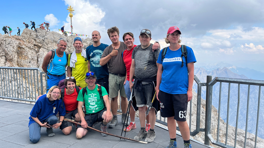 10 Menschen in Wanderklamotten lachen in die Kamera, im Hintergrund ist das Gipfelkreuz der Zugspitze zu sehen.
