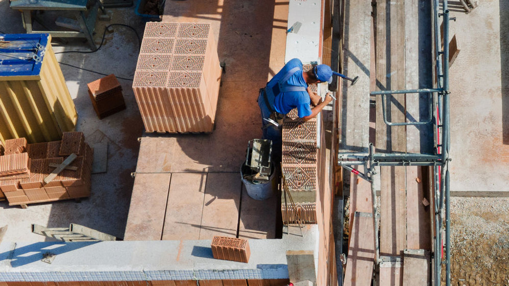 Ein Bauarbeiter mit blauem Helm und blauer Arbeitskleidung steht an der unfertigen Wand eines Neubaus. Hinter ihm liegen Ziegel bereit, die er auf der noch unfertigen Außenwand einfügt. 