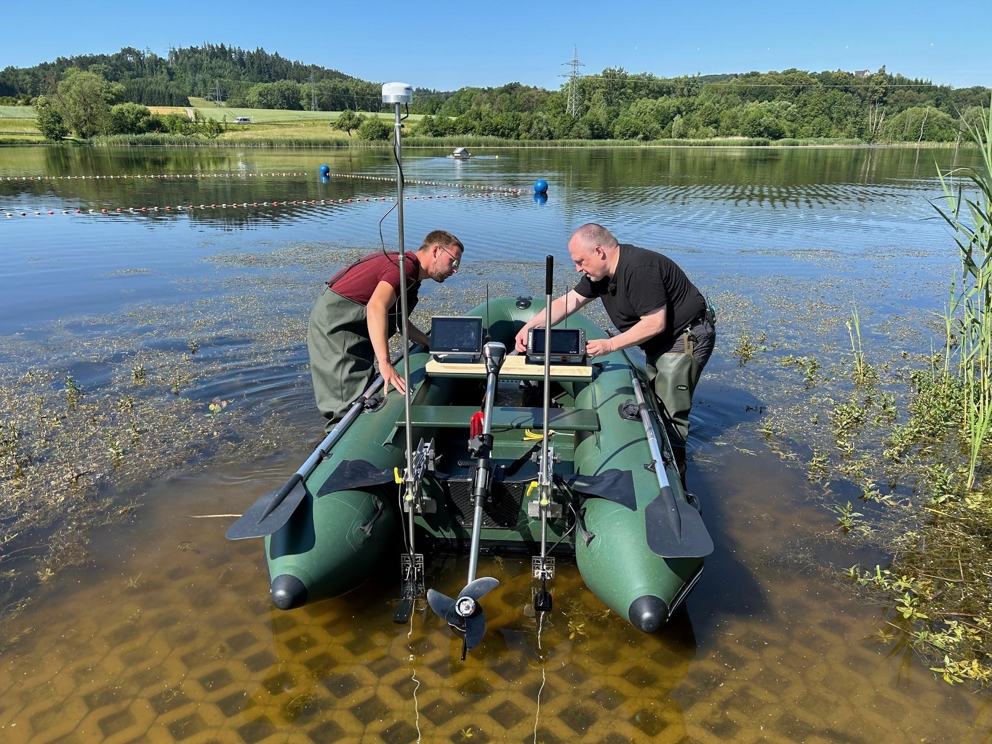 Zwei Männer stehen in Watthosen in einem See und befestigen technische Apparaturen an einem Boot.
