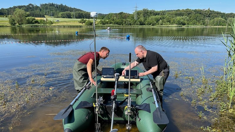 Zwei Männer stehen in Watthosen in einem See und befestigen technische Apparaturen an einem Boot. | Bild: BR / Andi Ebert Zwei Männer stehen in Watthosen in einem See und befestigen technische Apparaturen an einem Boot.