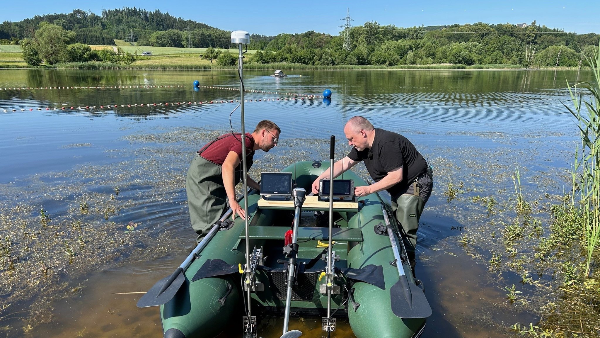 Zwei Männer stehen in Watthosen in einem See und befestigen technische Apparaturen an einem Boot.