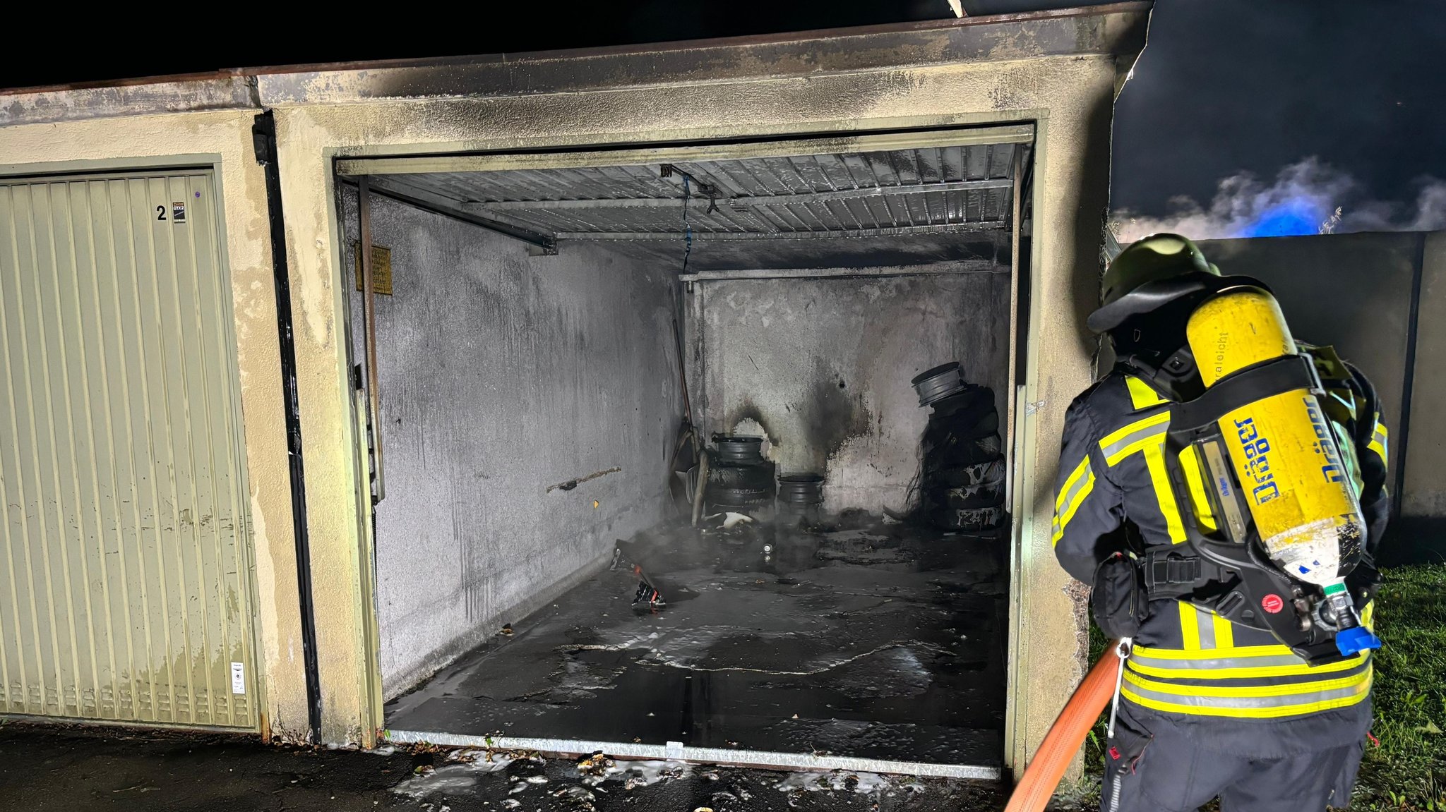 Ein Feuerwehrmann steht vor einer ausgebrannten Garage und schaut auf verkohlte Reifen im Inneren. Das Feuer hat er zuvor gelöscht, den Schlauch hält er noch in den Händen.