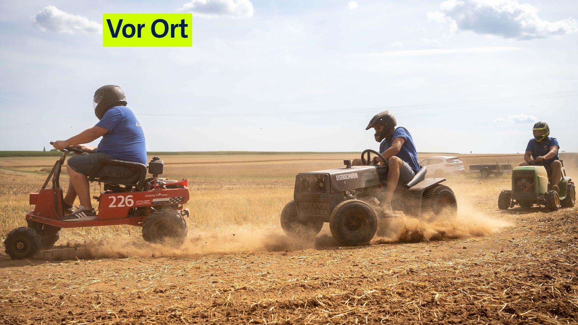 Ein Aufsitz-Rasenmäher fährt hinter einem anderen her, der Boden staubt. Die Fahrer tragen Helme.