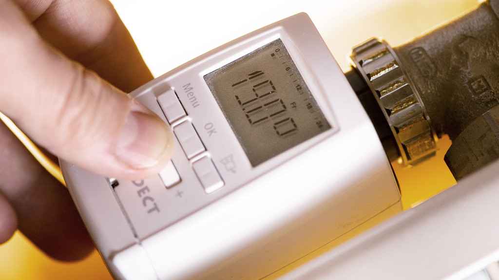 Eine Hand fasst an ein modernes Thermostat mit digitaler Anzeige an einer Heizung, wobei 19 Grad Celsius angezeigt werden.