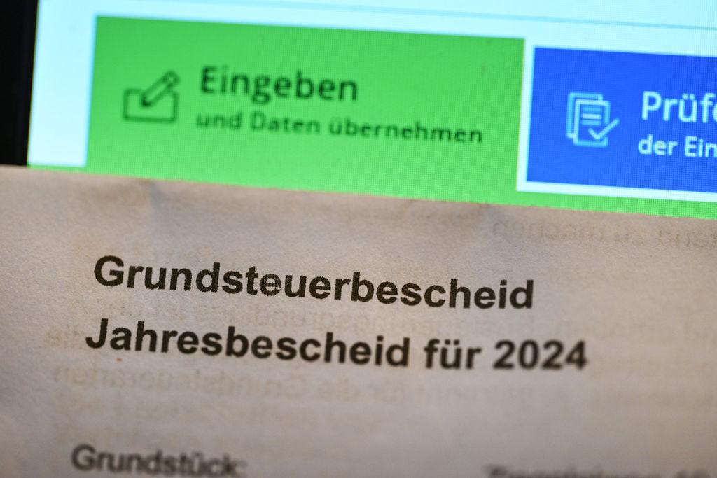 Ein Grundsteuerbescheid für 2024 wird vor einen Computerbildschirm gehalten, auf dem ein Formular der Steuerplattform Elster angezeigt wird