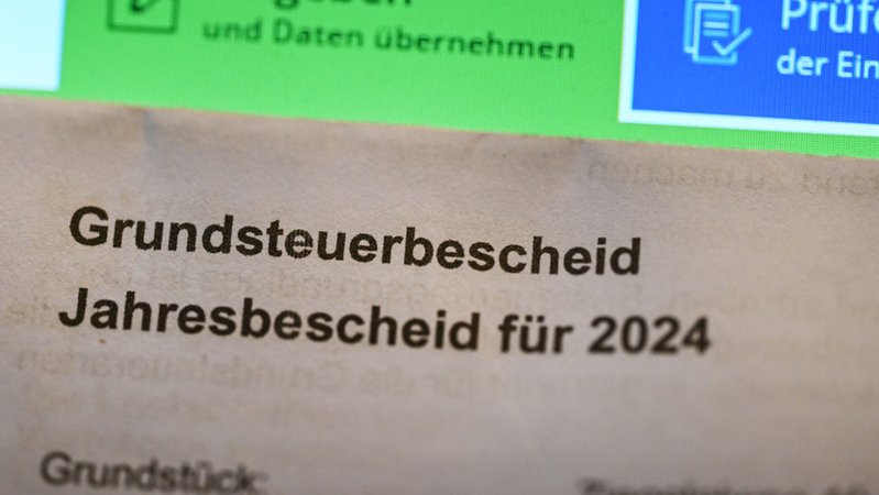 Ein Grundsteuerbescheid für 2024 wird vor einen Computerbildschirm gehalten, auf dem ein Formular der Steuerplattform Elster angezeigt wird | Bild: dpa-Bildfunk/Bernd Weißbrod Ein Grundsteuerbescheid für 2024 wird vor einen Computerbildschirm gehalten, auf dem ein Formular der Steuerplattform Elster angezeigt wird