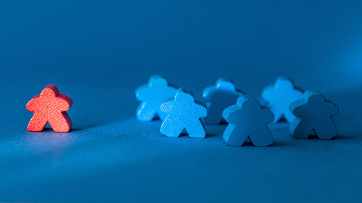 Fünf blaue Spielfiguren stehen in einem Kreis eine rote Spielfigur steht etwas abseits in der Nähe einer nicht sichtbaren Lichtquelle.