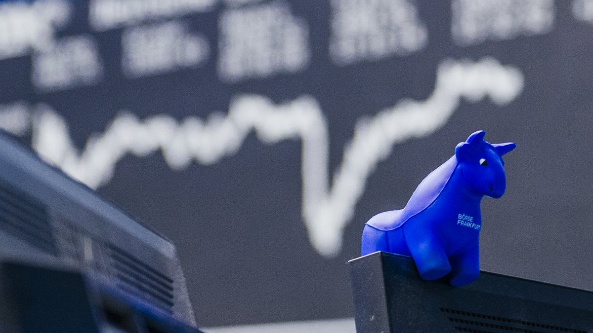 Ein blauer Stier aus Gummi sitzt auf der Kante eines Bildschirms, im Hintergrund die Kurstafel der Börse