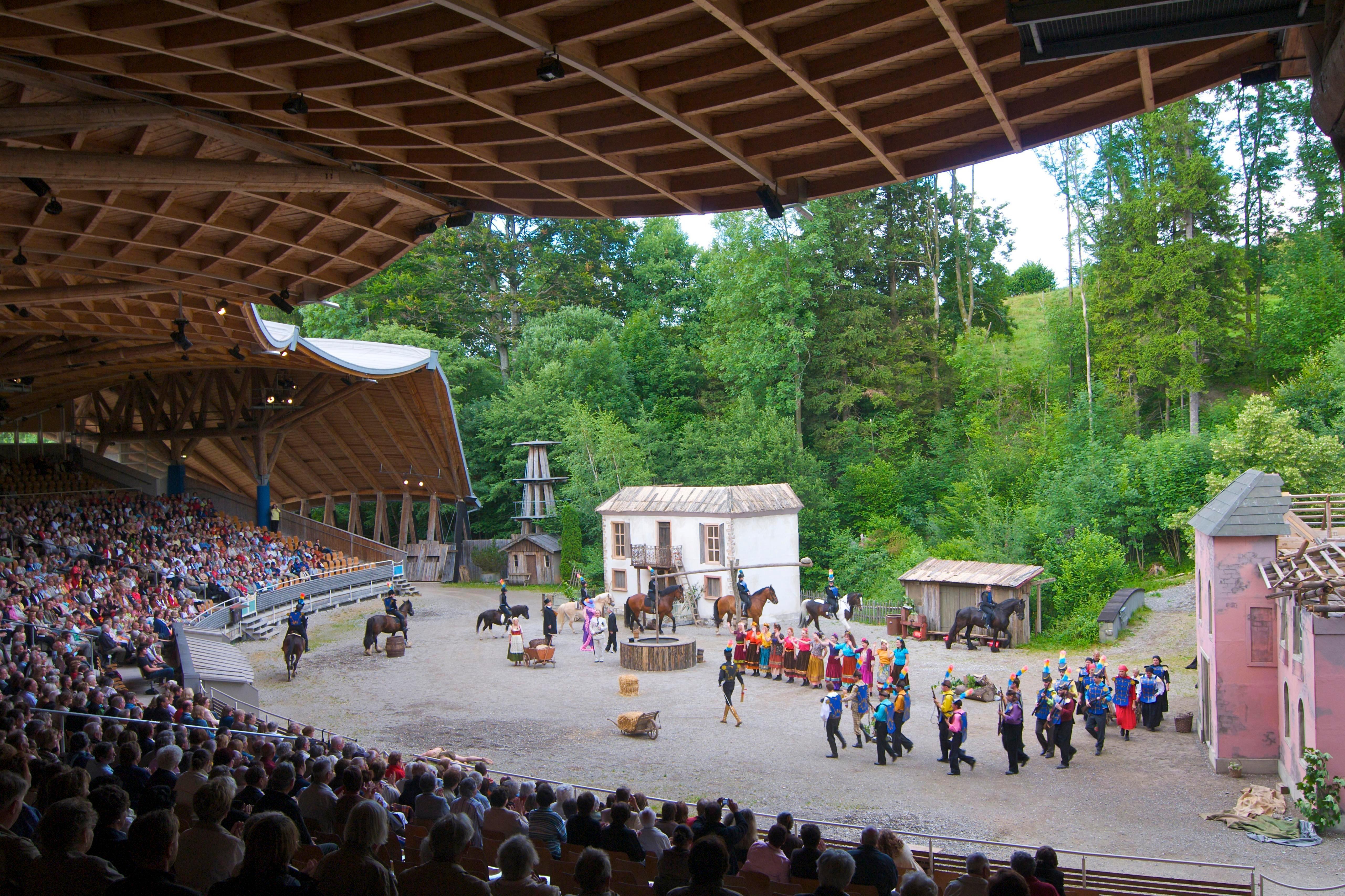 Blick von der Tribüne in Altusried, die von einer geschwungenen Holzkonstruktion überdacht ist, auf die Freilichtbühne, auf der Kulissen stehen und bunt gekleidete Schauspielerinnen und Schauspieler, teils auf Pferden, zu sehen sind. Bis zu 2500 Besucher finden bei Aufführungen unter dem freischwebenden Tribünendach der Allgäuer Freilichtspiele Altusried Platz (Symbolbild).