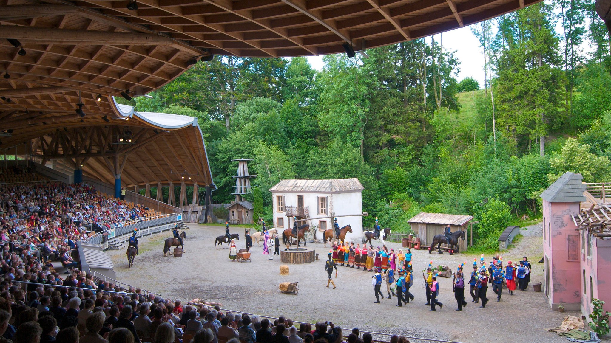 Blick von der Tribüne in Altusried, die von einer geschwungenen Holzkonstruktion überdacht ist, auf die Freilichtbühne, auf der Kulissen stehen und bunt gekleidete Schauspielerinnen und Schauspieler, teils auf Pferden, zu sehen sind. Bis zu 2500 Besucher finden bei Aufführungen unter dem freischwebenden Tribünendach der Allgäuer Freilichtspiele Altusried Platz (Symbolbild).