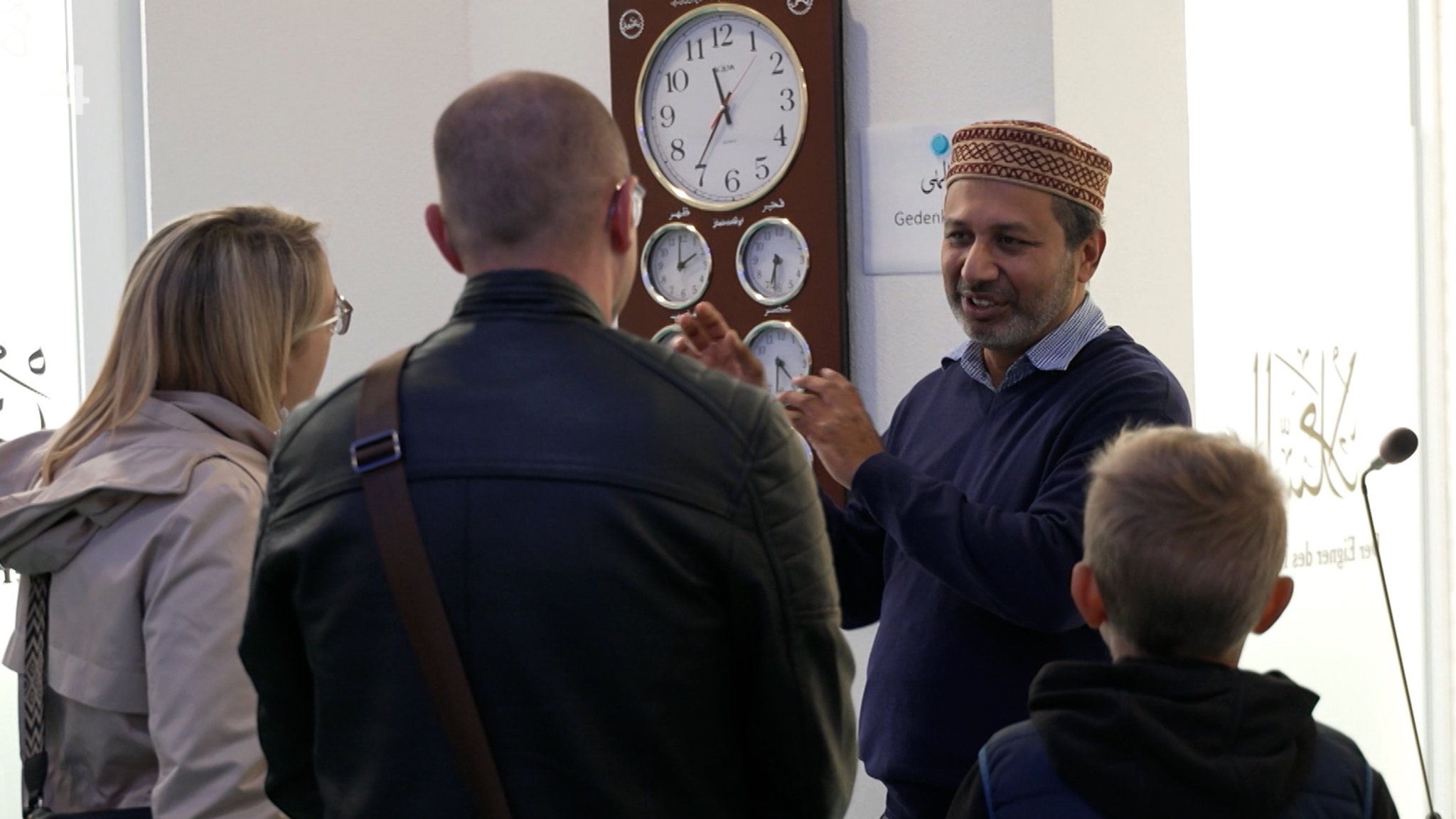 Eine Person steht neben einer Wand mit vielen Uhren und spricht zu drei Personen, die nur von hinten zu sehen sind. Am "Tag der offenen Moschee" hat auch die Baitunnaseer Moschee in Augsburg-Oberhausen für Besucherinnen und Besucher geöffnet.