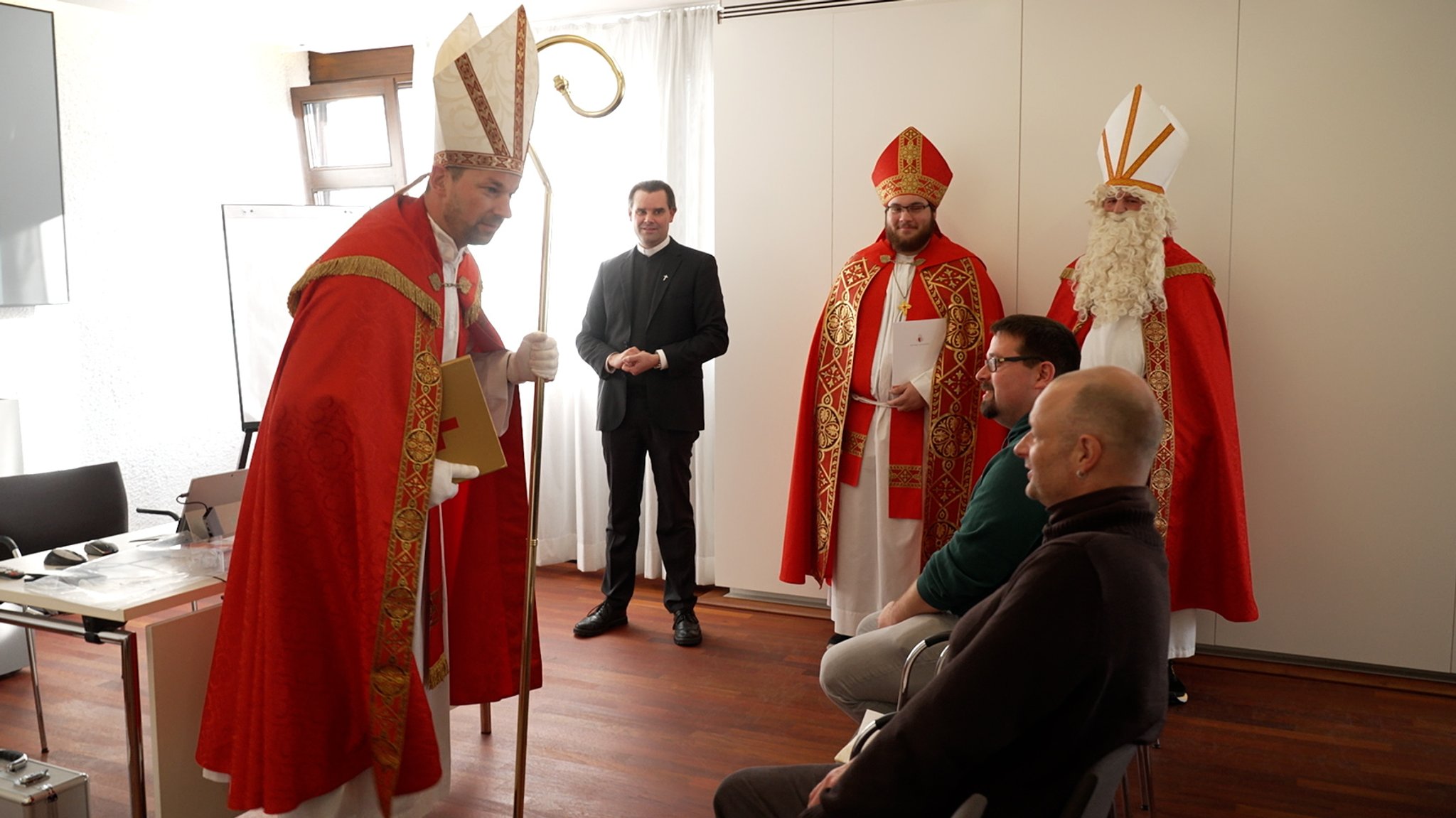 Ein als Bischof Nikolaus verkleideter Mann steht vor zwei sitzenden Männern in einem Seminarraum. Beim "Nikolaus-Zertifikatskurs" dürfen sich die Teilnehmer in einem Rollenspiel als Heiliger Nikolaus ausprobieren.