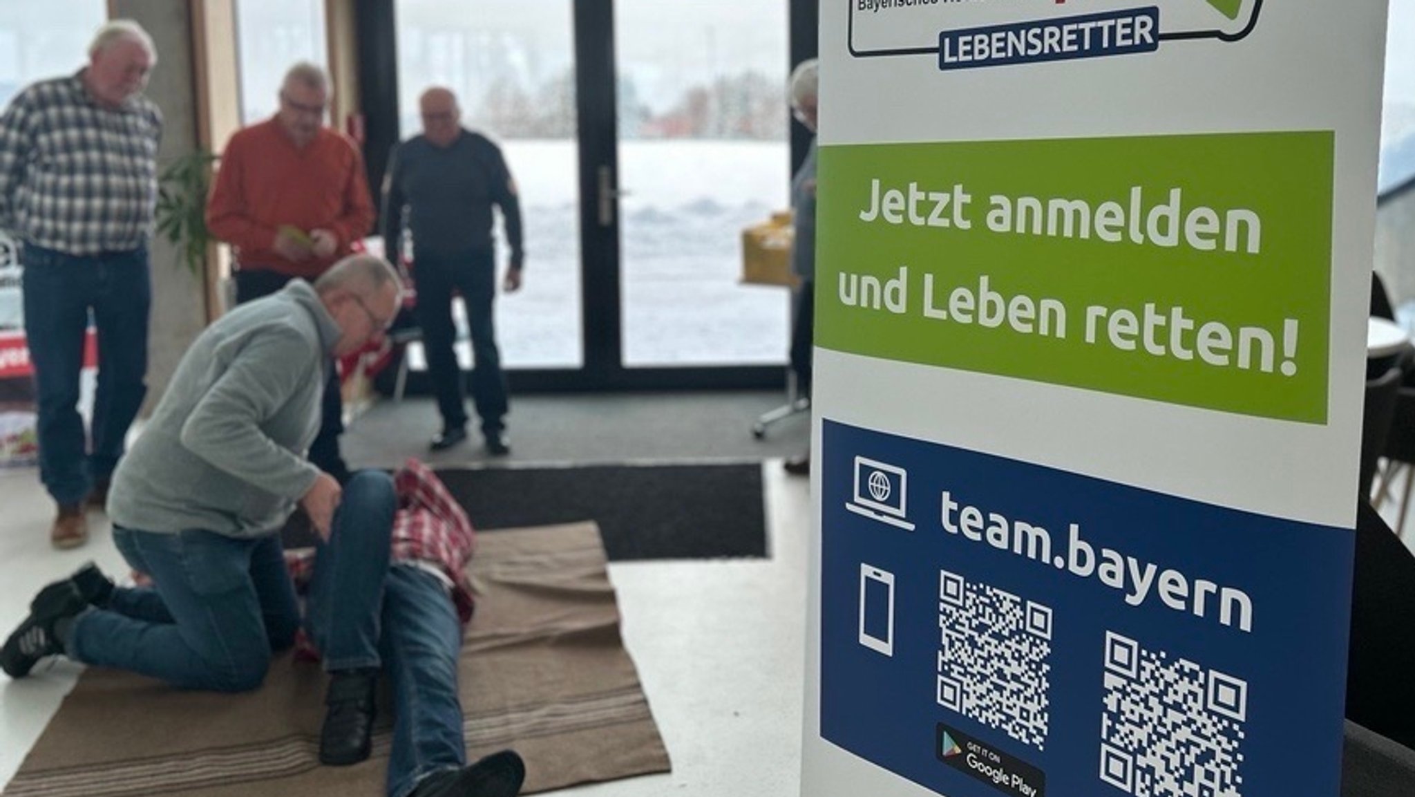 "Team Bayern Lebensretter" steht auf einem Roll-Up. Im Hintergrund kniet ein Mann über einer anderen Person. Drei weitere Männer sehen zu. 