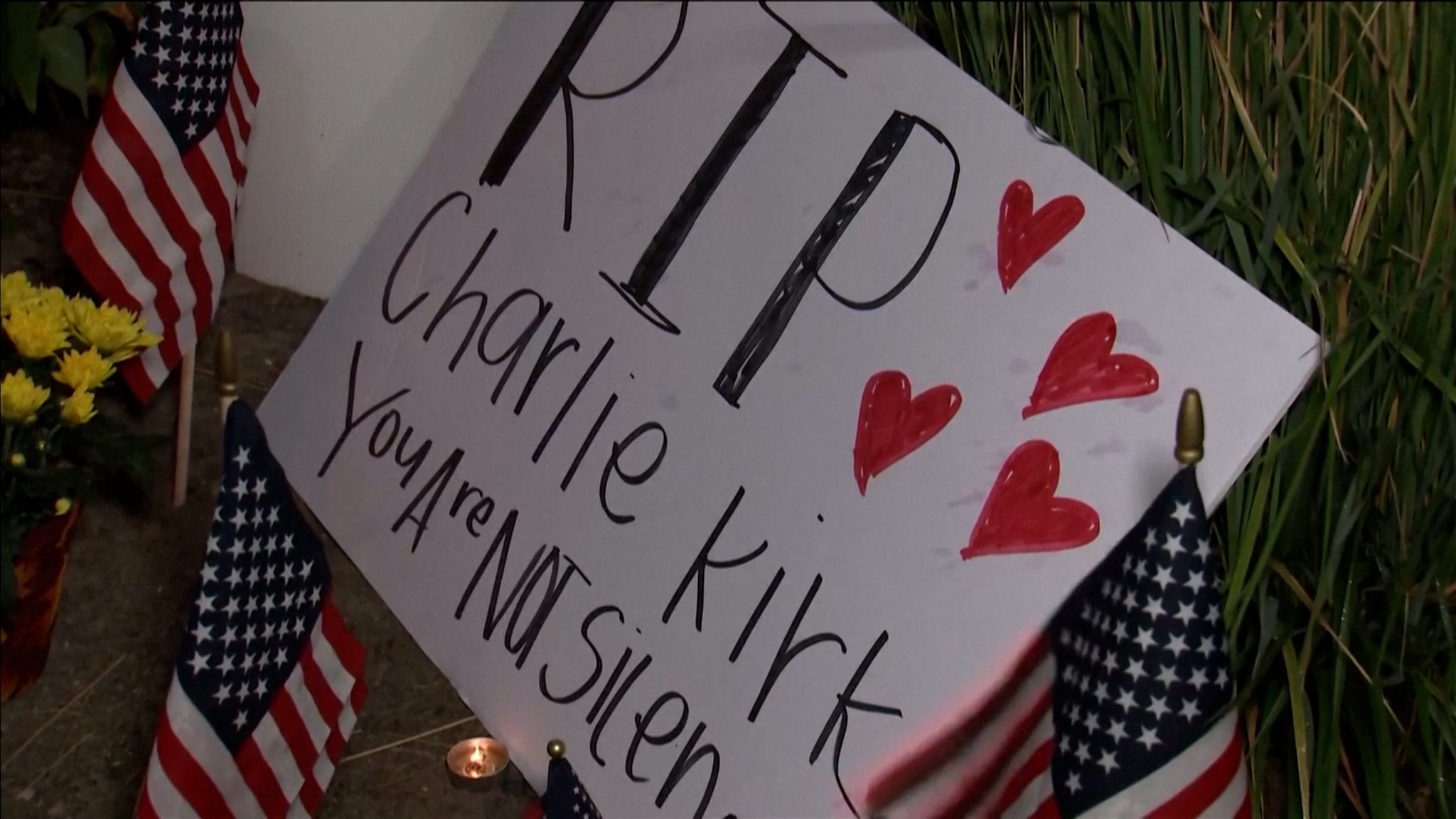 Ein Schild mit der Aufschrift "RIP Charlie Kirk" und roten Herzen liegt zwischen US-Flaggen und Teelichtern auf dem Boden