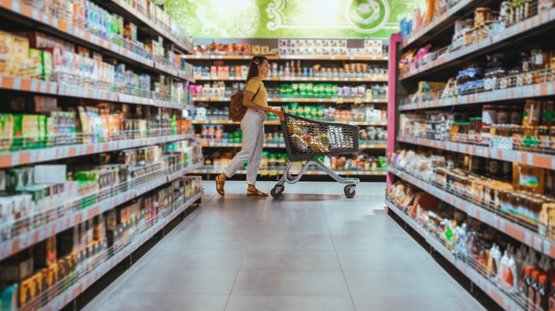 Symbolbild: Supermarktregale, im Hintergrund eine Frau mit Einkaufswagen | Bild: stock.adobe.com/phpetrunina14 Symbolbild: Supermarktregale, im Hintergrund eine Frau mit Einkaufswagen