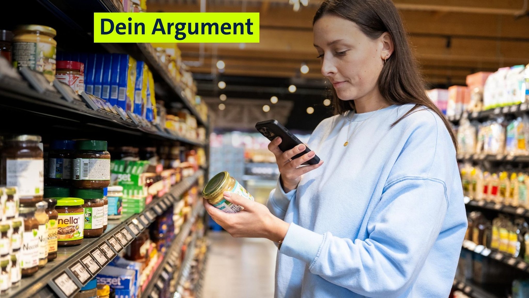 Eine Frau ist beim Einkauf in einem Supermarkt zu sehen. Sie hält ein Glas mit Lebensmitteln in der einen Hand und ein Smartphone in der anderen. (Symbolbild) | Bild: picture alliance / dpa-tmn | Benjamin Nolte Eine Frau ist beim Einkauf in einem Supermarkt zu sehen. Sie hält ein Glas mit Lebensmitteln in der einen Hand und ein Smartphone in der anderen. (Symbolbild)