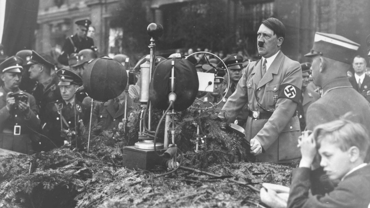 Diktator Adolf Hitler am 1. Mai 1934 vor aufgebauten Mikrofonen, hinter ihm eine Menschenmenge, die meisten davon Männer in Uniformen.   