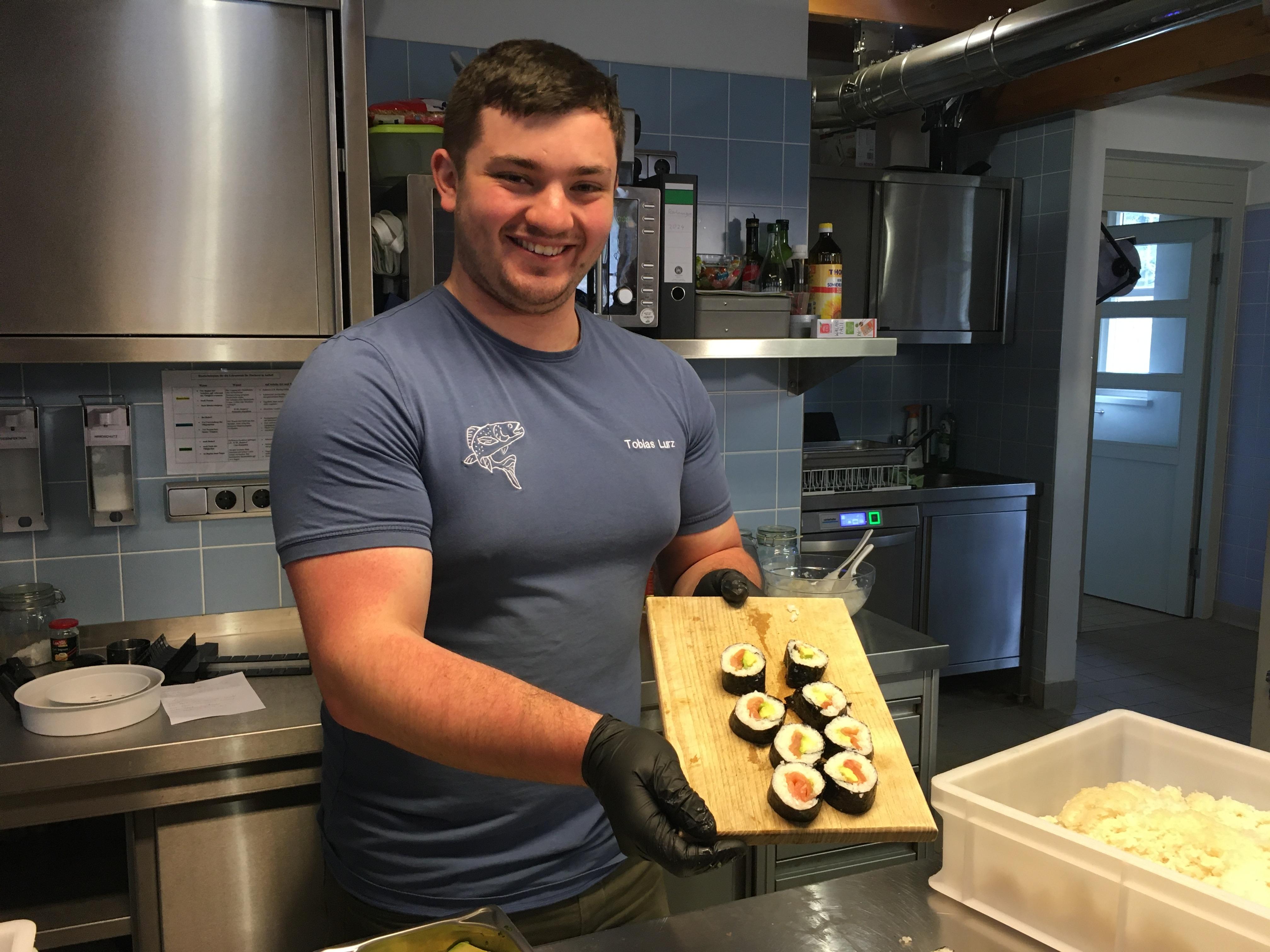 Ein Mann steht in einer Küche und hält grinsend eine Holzplatte in der Hand, auf der acht Sushi-Rollen liegen. 