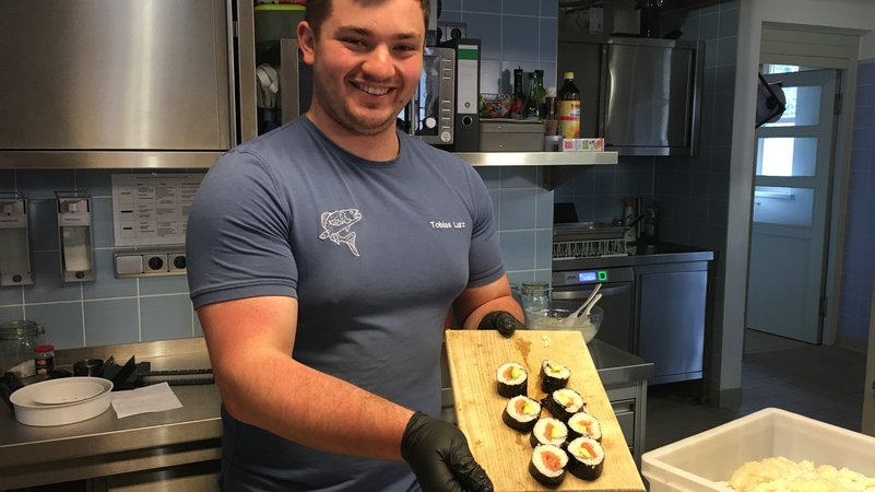 Ein Mann steht in einer Küche und hält grinsend eine Holzplatte in der Hand, auf der acht Sushi-Rollen liegen. 