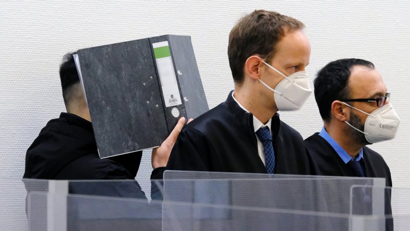 Ein Angeklagter verbirgt im Sitzungssaal des Landgerichts Ingolstadt sein Gesicht hinter einem Aktenordner. Davor stehen seine Verteidiger Andreas Ruch und Adam Ahmed (r). 