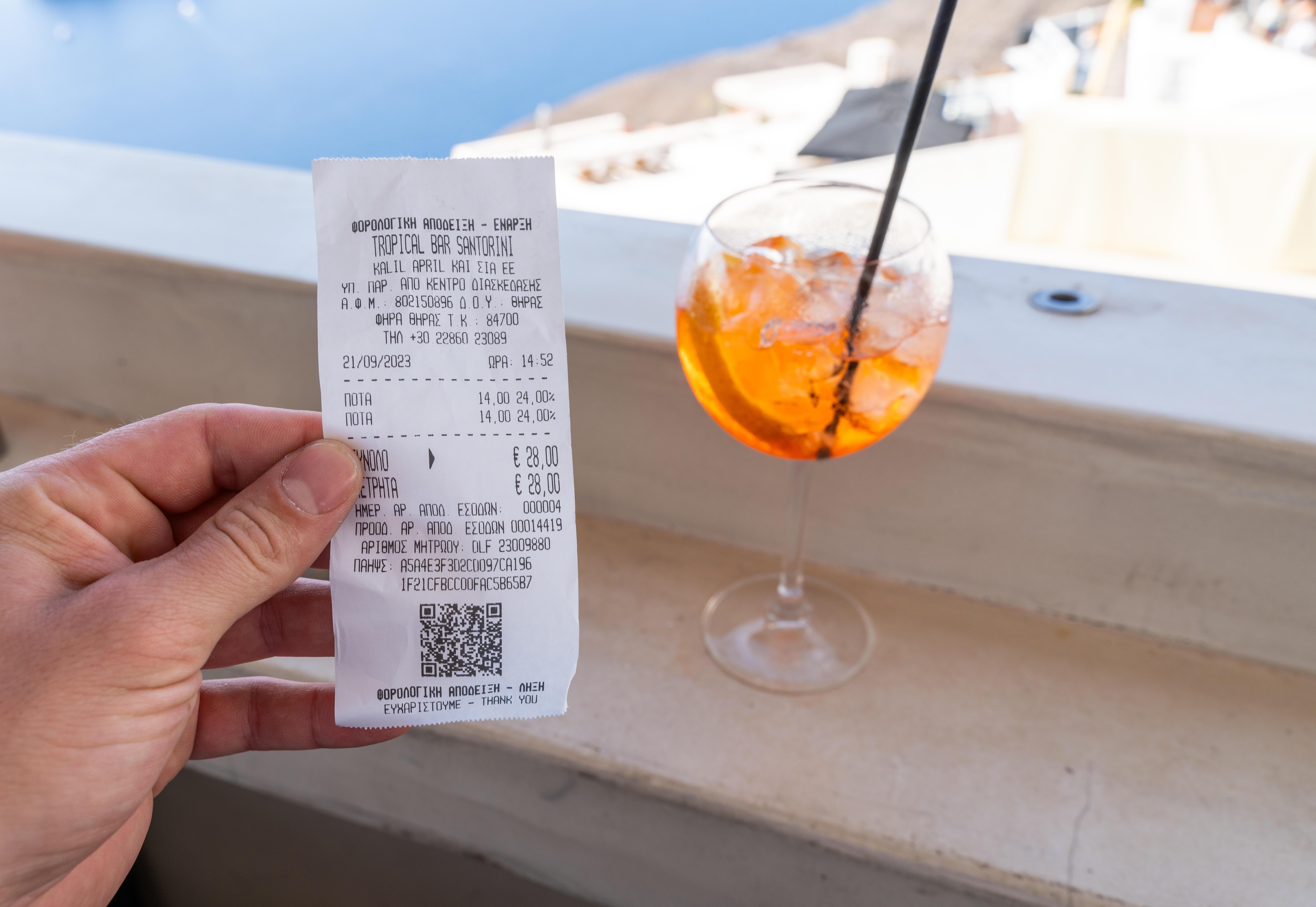 Ein Tourist hält eine Rechnung in einer Bar vor ein alkoholisches Getränk auf Santorini in Griechenland