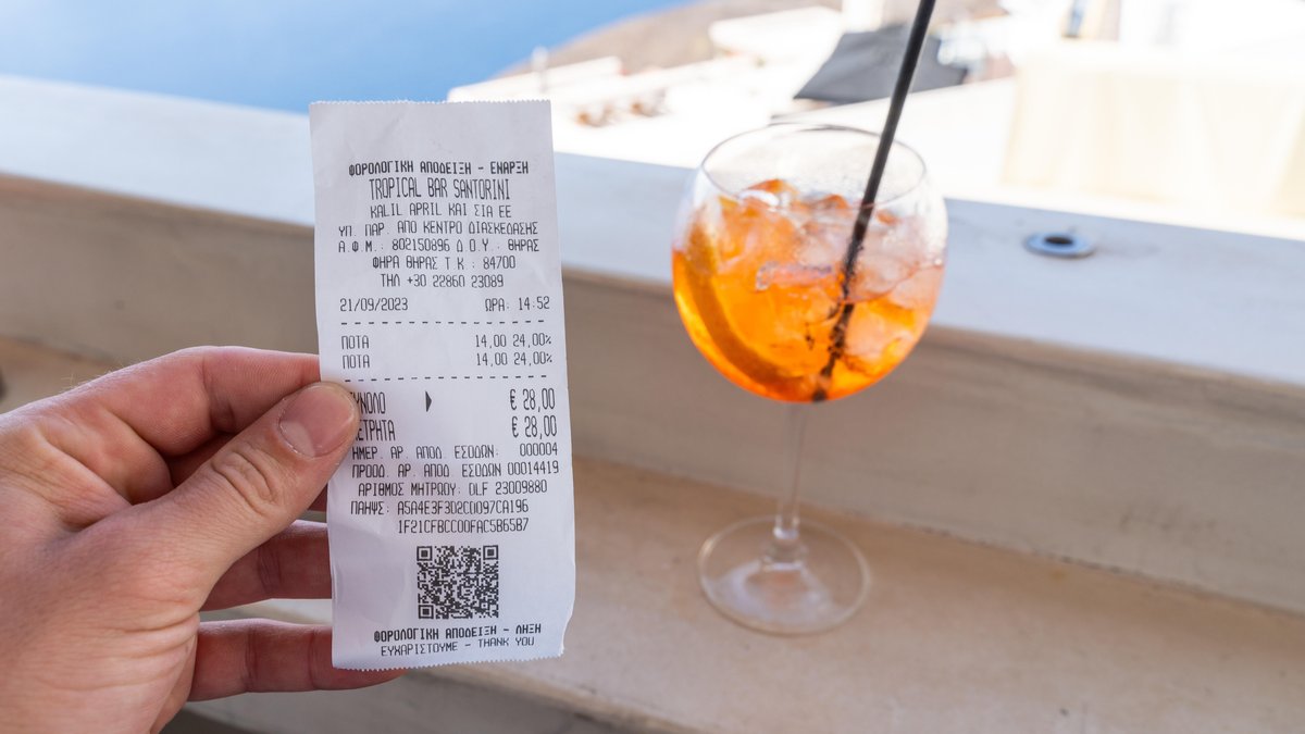 Ein Tourist hält eine Rechnung in einer Bar vor ein alkoholisches Getränk auf Santorini in Griechenland