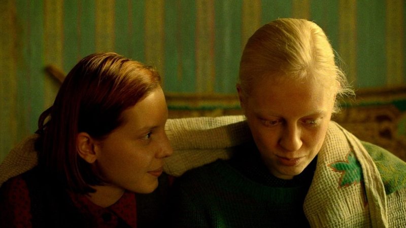 Zwei junge Frauen sitzen zusammen unter einer Decke in einem Raum in mildem Licht (Filmszene aus: "Bohnenstange")