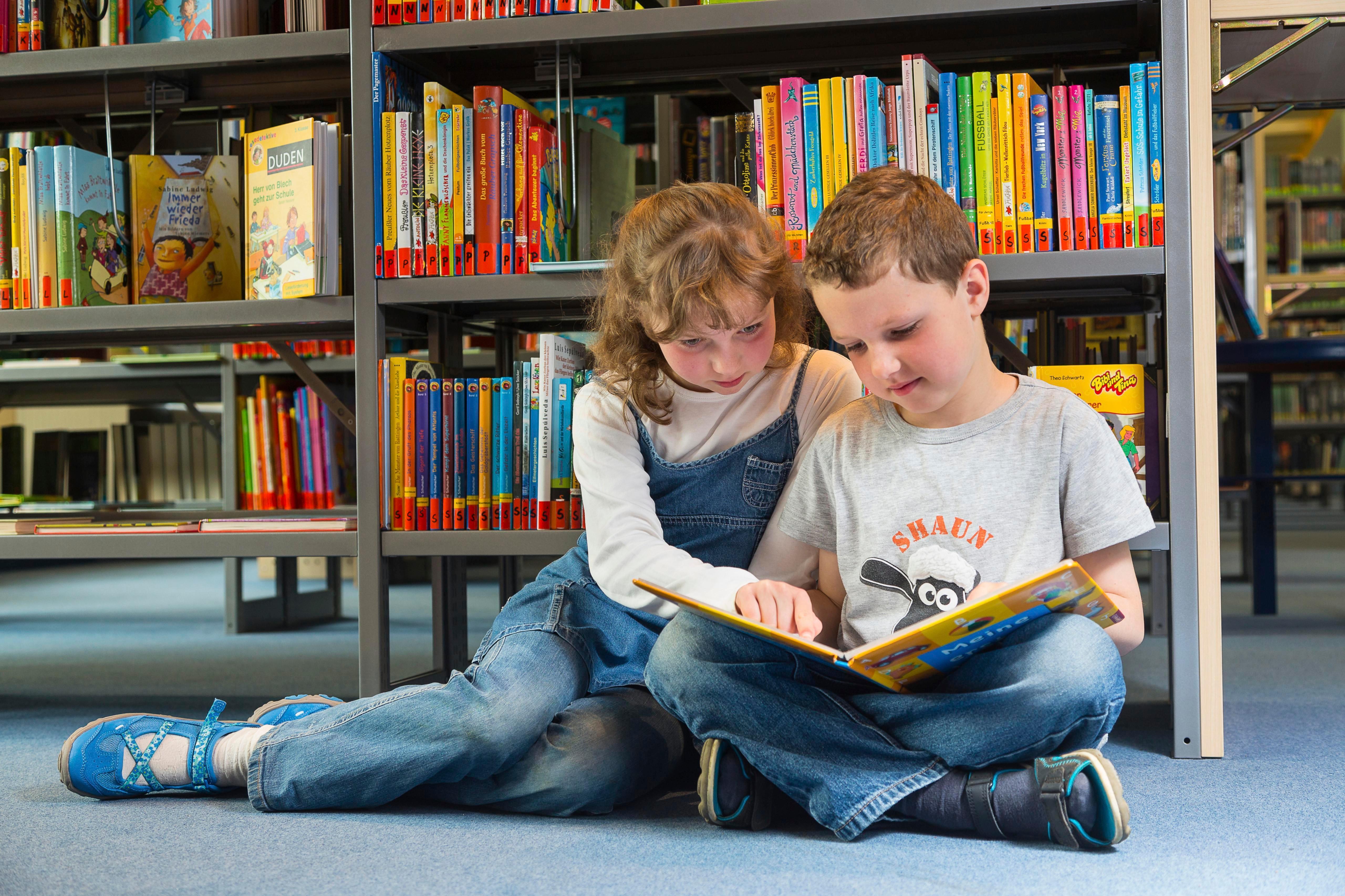 Zwei Kinder sitzen in einer Bücherei auf dem Boden und lesen gemeinsam in einem Kinderbuch  | Bild:picture alliance/imageBROKER/Gabriele Hanke