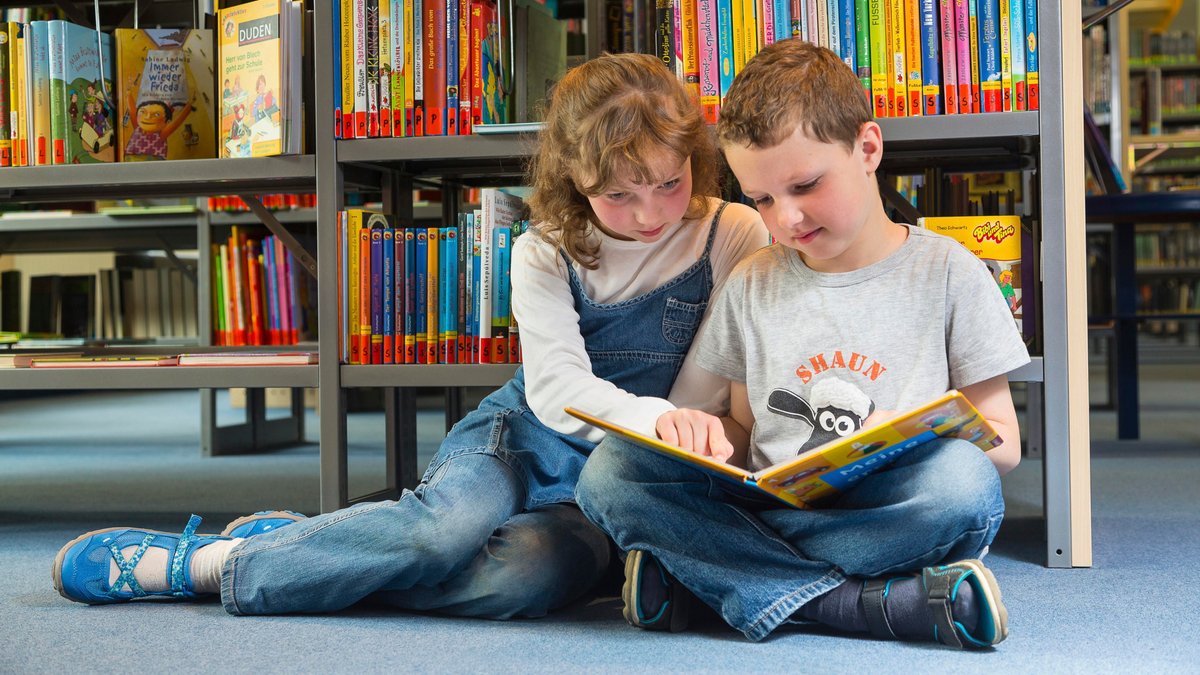 Zwei Kinder sitzen in einer Bücherei auf dem Boden und lesen gemeinsam in einem Kinderbuch 