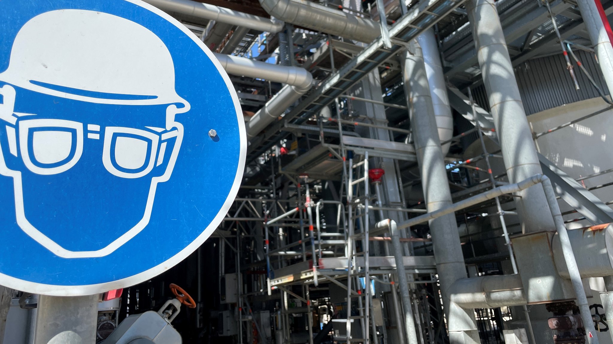 Eine Baustelle auf dem Bayernoil-Gelände in Neustadt, im Vordergrund ein blaues Schild, das auf die Pflicht zum Tragen einer Schutzausrüstung hinweist.