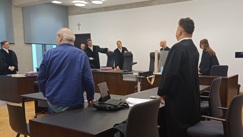 Ein Angeklagter in blauem Kittel in einem Gerichtssaal neben seinem Anwalt.