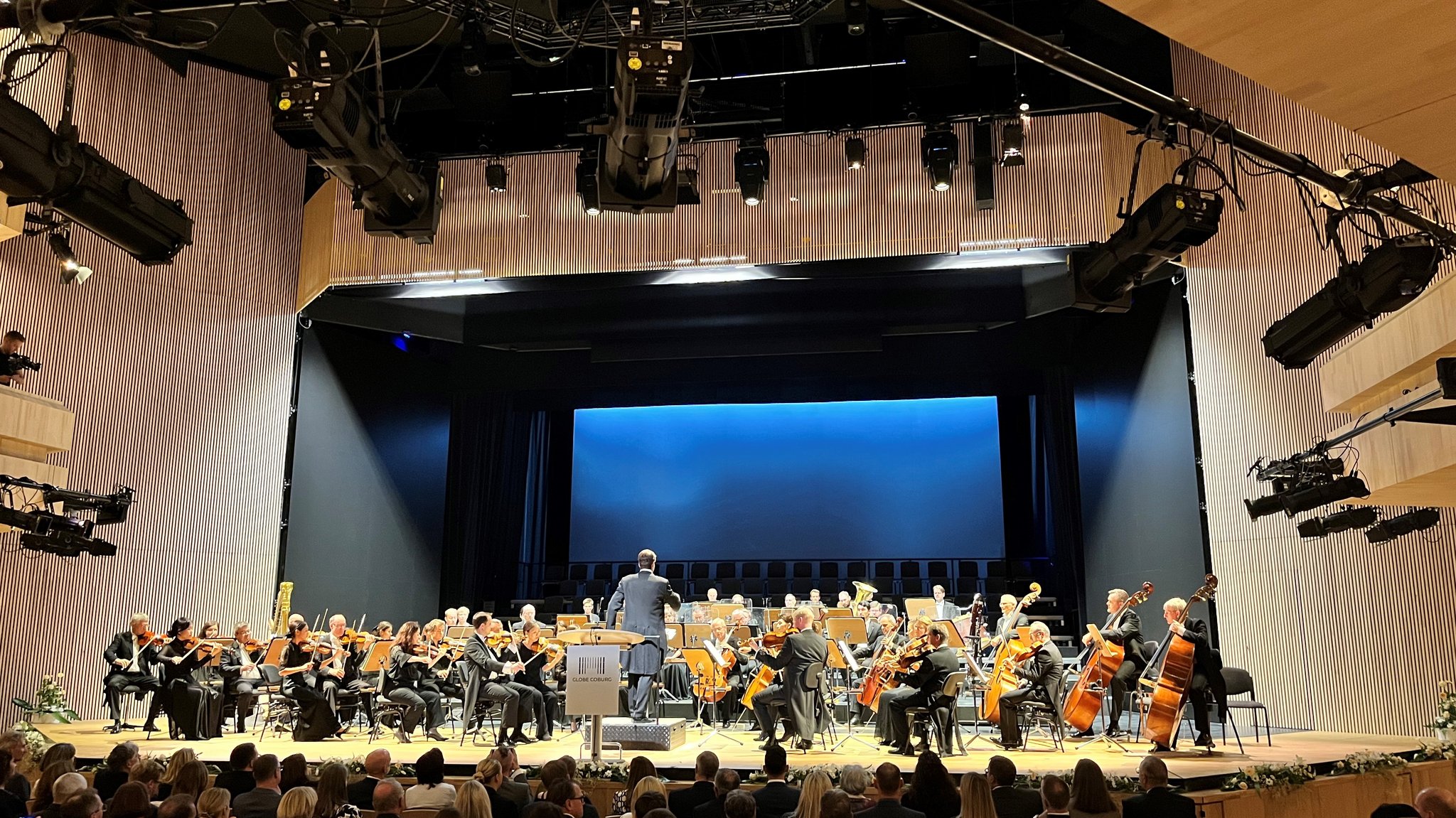 Ein Orchester spielt auf der Bühne, davor sitzen viel Publikum, das zuhört.  