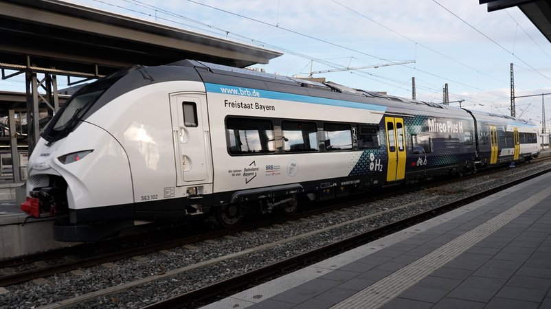 Der Wasserstoffzug fährt in den Augsburger Hauptbahnhof ein. Der Zug ist weiß mit einem dunkelblauen Streifen in der Mitte und gelben Türen, die Scheinwerfer erinnern an Katzenaugen. | Bild: BR/Johannes Hofelich Der Wasserstoffzug fährt in den Augsburger Hauptbahnhof ein. Der Zug ist weiß mit einem dunkelblauen Streifen in der Mitte und gelben Türen, die Scheinwerfer erinnern an Katzenaugen.