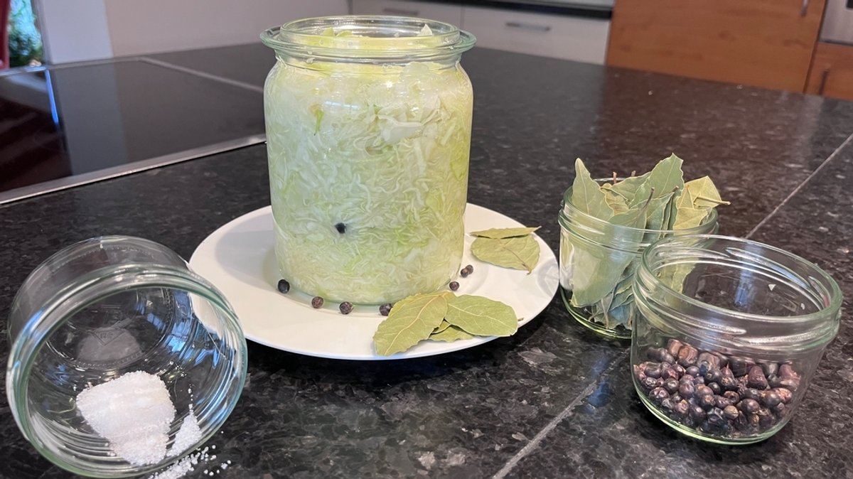 Sauerkraut in einem Glas, dazu die Zutaten Salz, Lorbeer und Wacholder