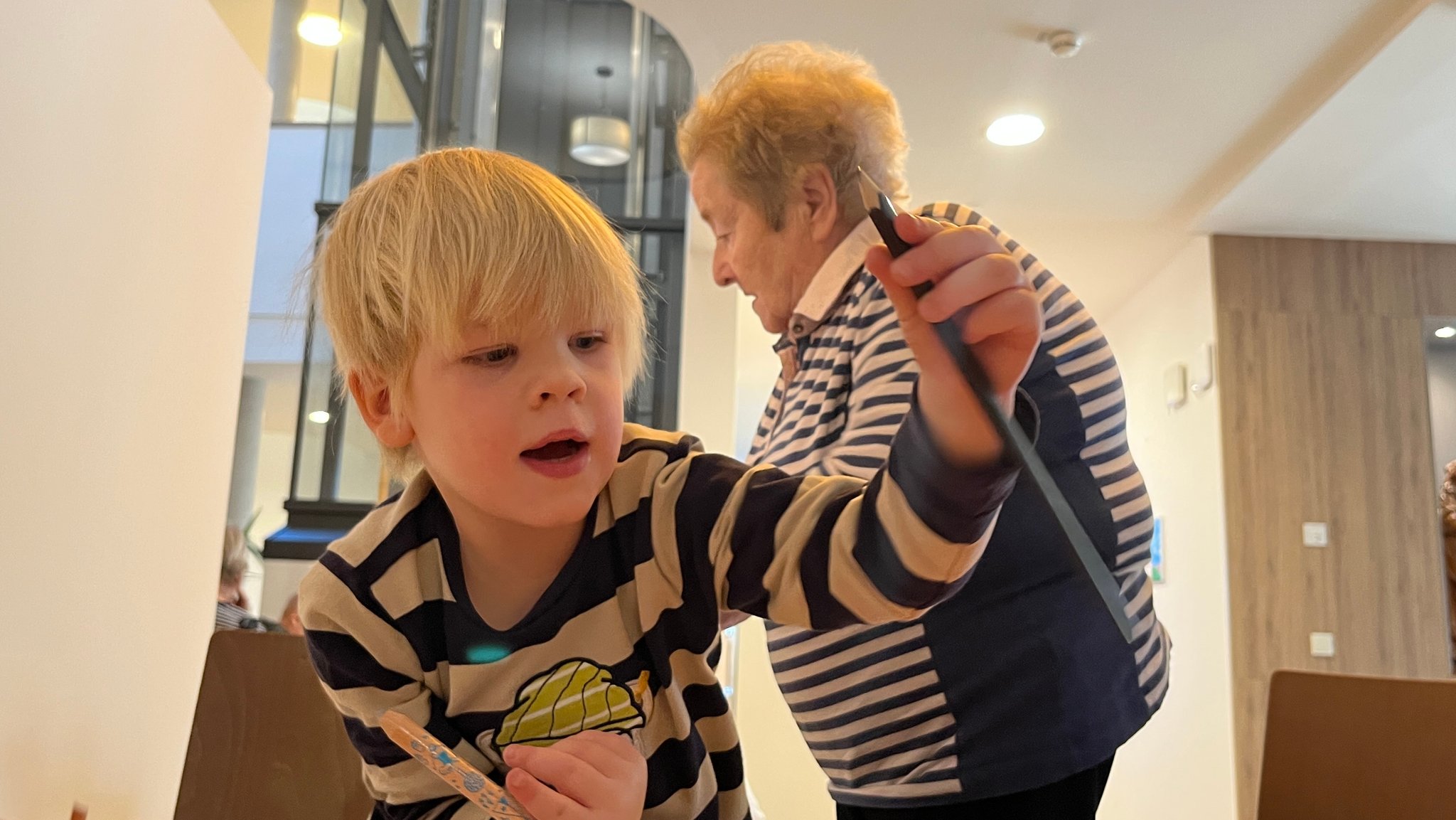 Ein blonder junge hebt einen Stift hoch und schaut nach unten auf sein Bild. Hinter ihm steht eine Seniorin.