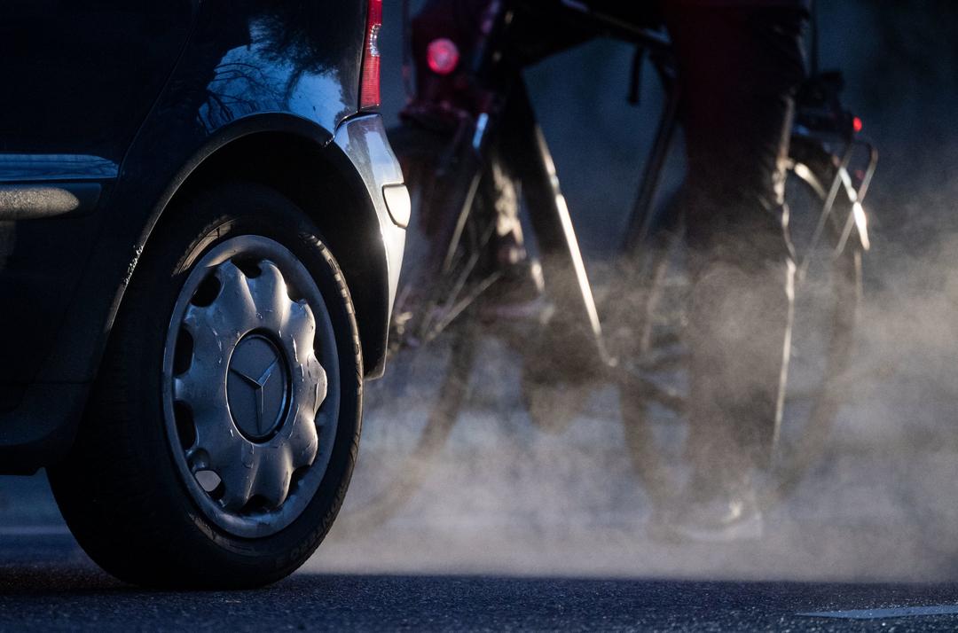 Ein Radfahrer steht neben einem Auto, dessen Abgase in der kalten Morgenluft sichtbar werden. 
