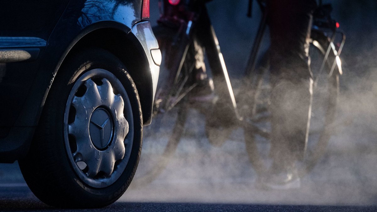 Ein Radfahrer steht neben einem Auto, dessen Abgase in der kalten Morgenluft sichtbar werden. 