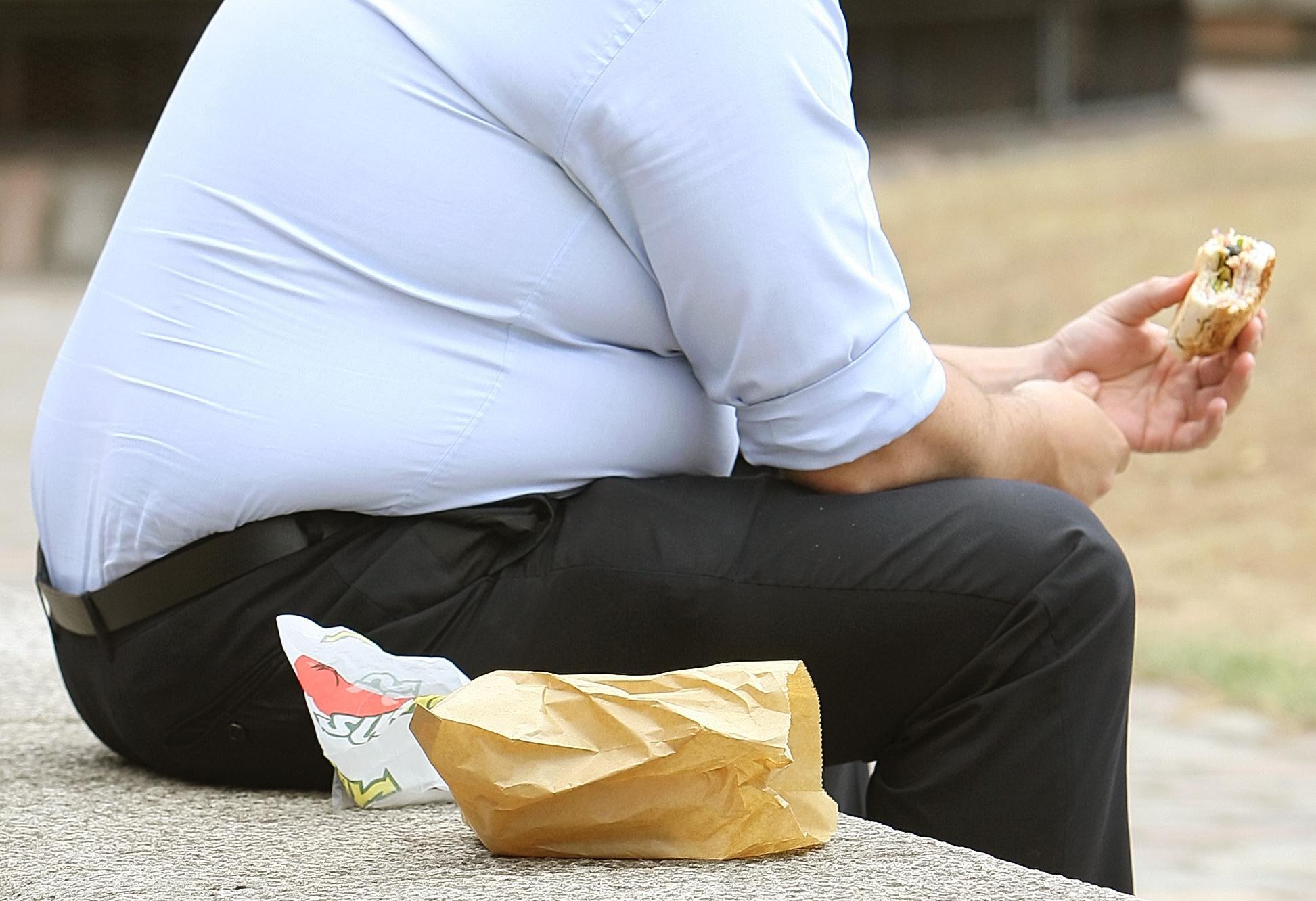 Ein übergewichtiger Mann sitzt auf einer Mauer und hält einen Burger in der Hand.