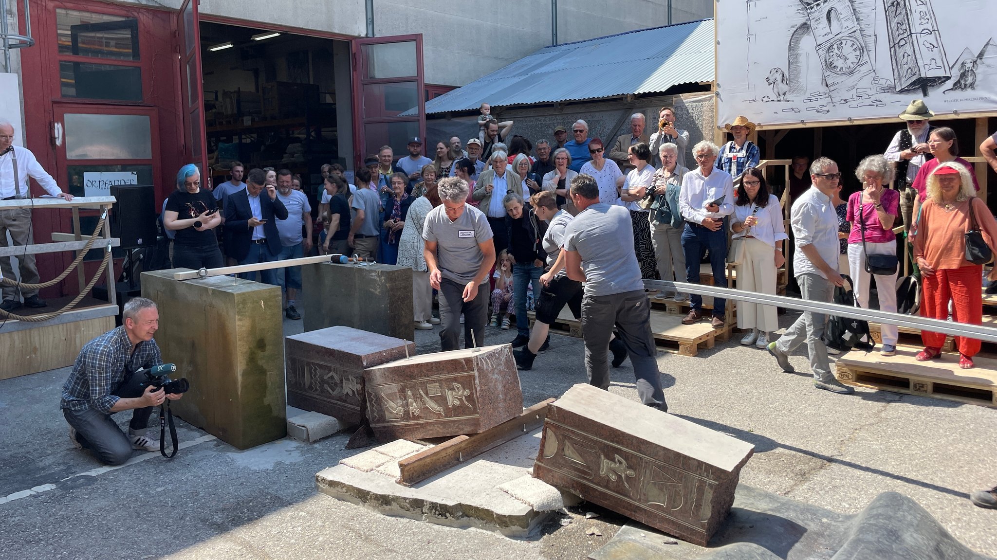 In mehrere Teile zerbrochener Obelisk aus rötlichem Granit am Boden vor der Werkshalle der Pfanner GmbH