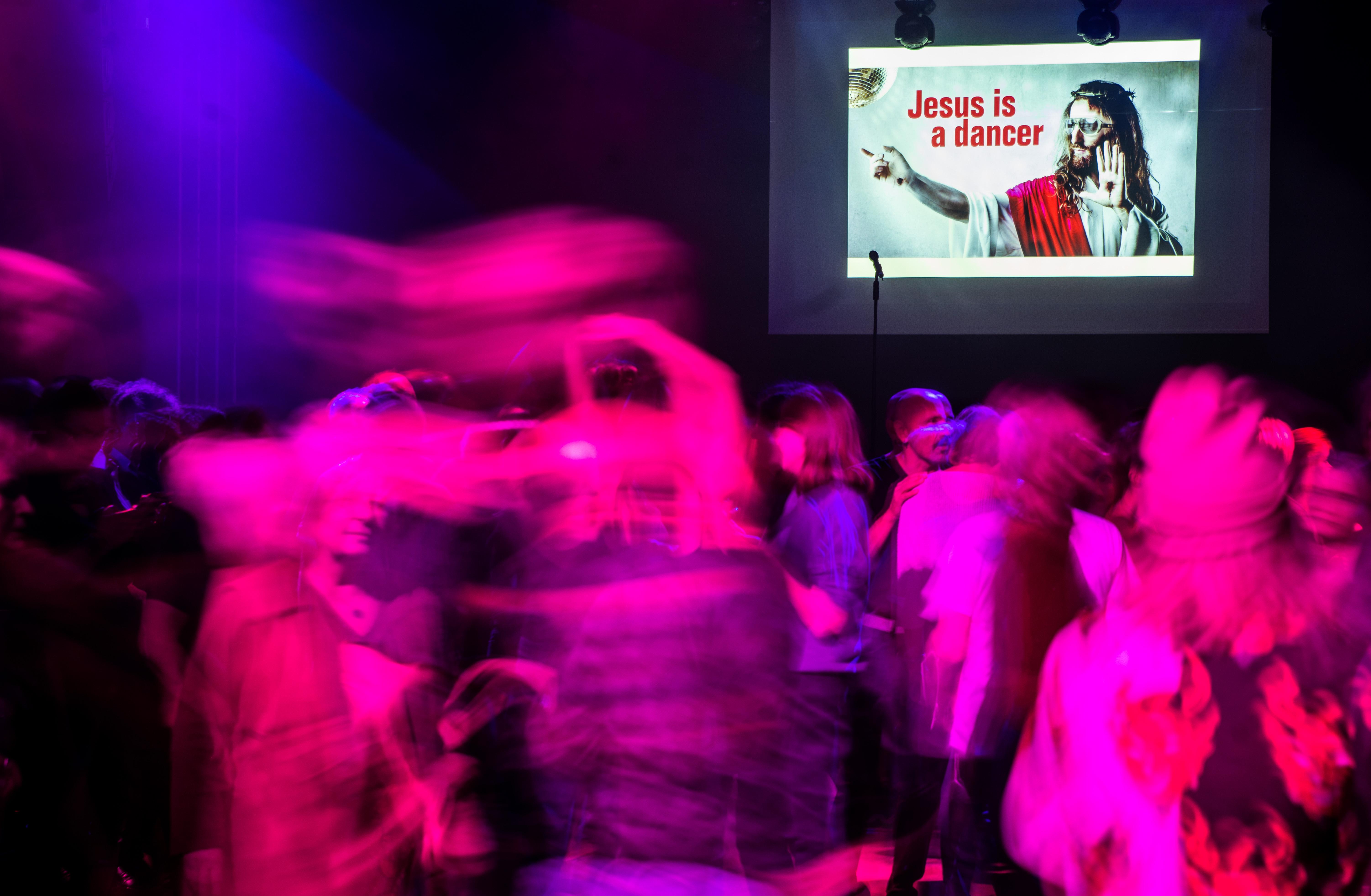 Club-Szene mit vielen Tanzenden. An der Wand ist ein Bild mit der Aufschrift "Jesus is a dancer". 