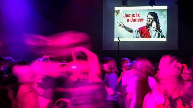Club-Szene mit vielen Tanzenden. An der Wand ist ein Bild mit der Aufschrift "Jesus is a dancer".  | Bild:Picture Alliance/dpa/Christoph Schmidt