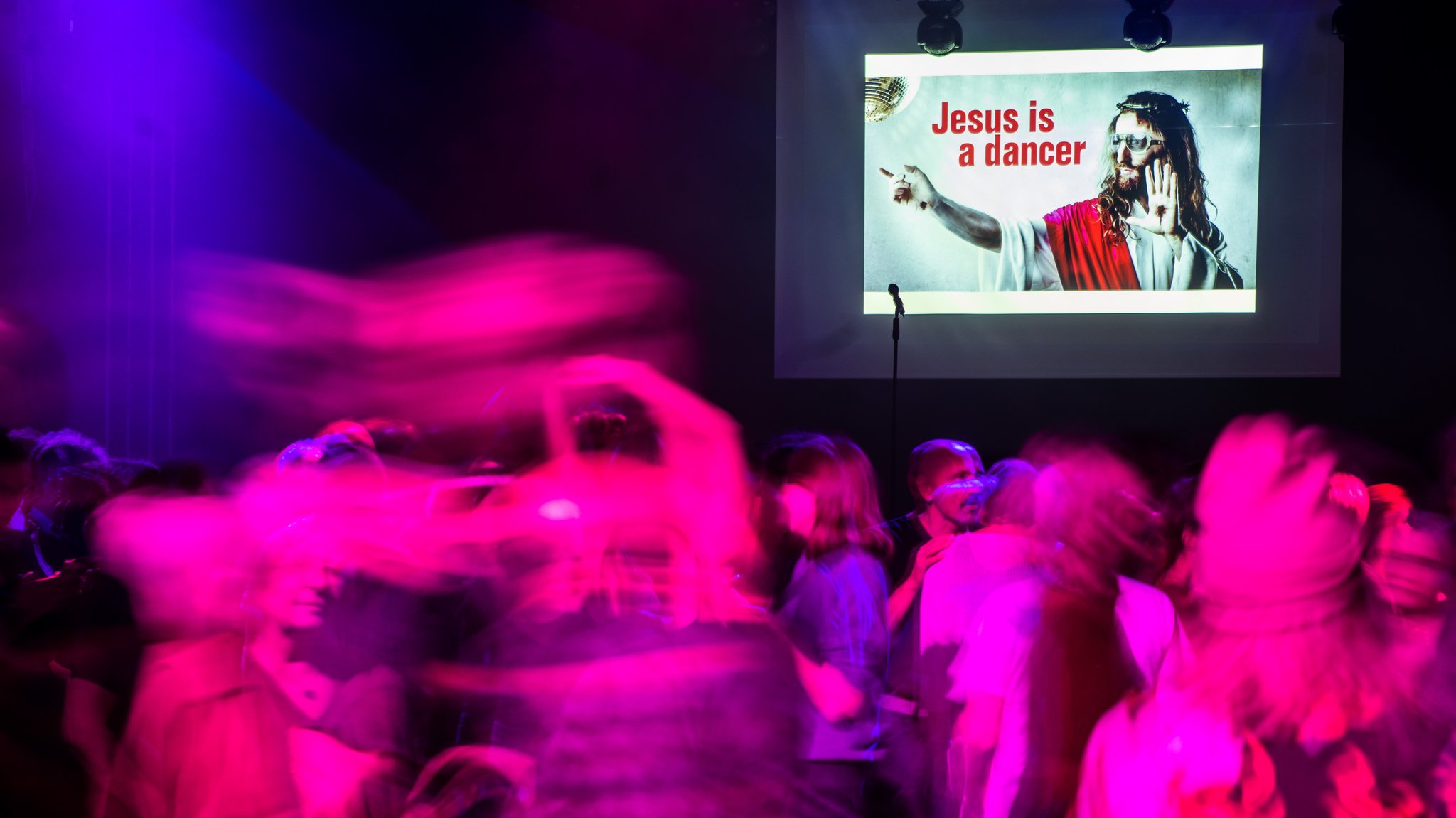 Club-Szene mit vielen Tanzenden. An der Wand ist ein Bild mit der Aufschrift "Jesus is a dancer". 