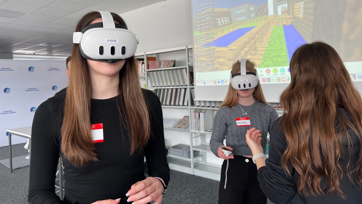 Zwei junge Frauen stehen mit einer weißen VR-Brille vor einem Bildschirm. Eine andere Frau erklärt, einer Schülerin mit VR-Brille etwas. Auf dem Bildschirm im Hintergrund sieht man Gebäude in einer virtuellen Welt, welche die jungen Frauen mit ihren VR-Brillen sehen.