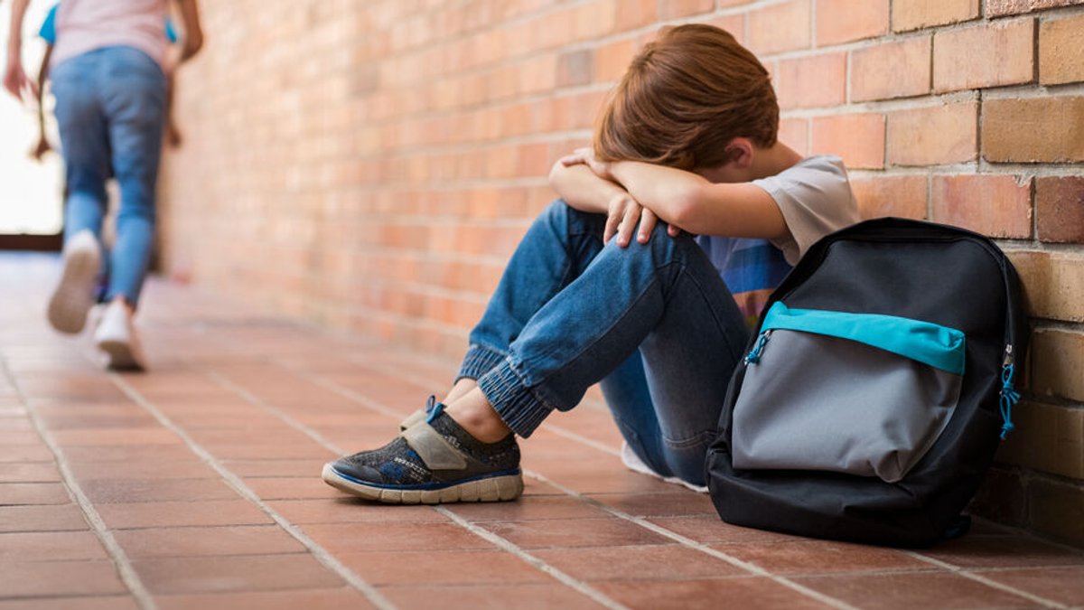 Ein Junge sitzt auf dem Flur in seiner Schule mit dem Rücken zur Wand und legt seinen Kopf zwischen seine Arme. Neben ihm liegt sein Schulranzen, Kinder laufen durch den Flur nach draußen.