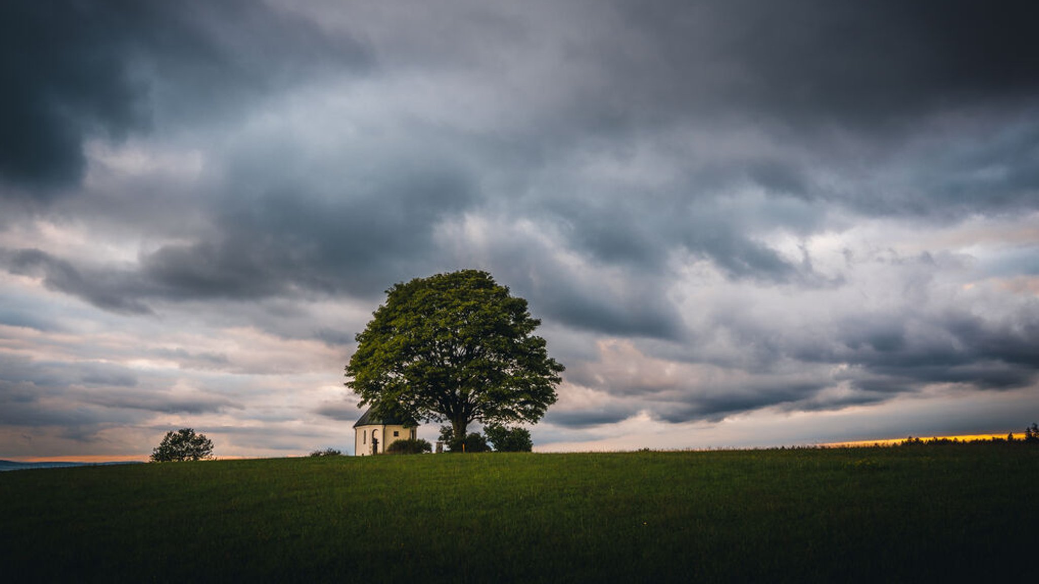 Dramatische Wolken in der Abenddämmerung über einer Wiese mit Kapelle und Baum.