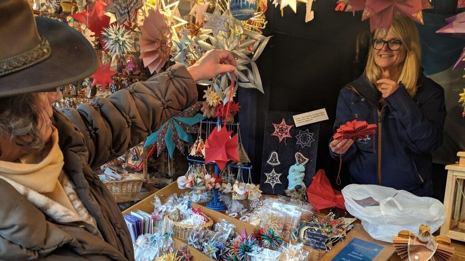 Eine Frau in einem Stand voller Bastelsterne zeigt lachend einen Papierstern. 