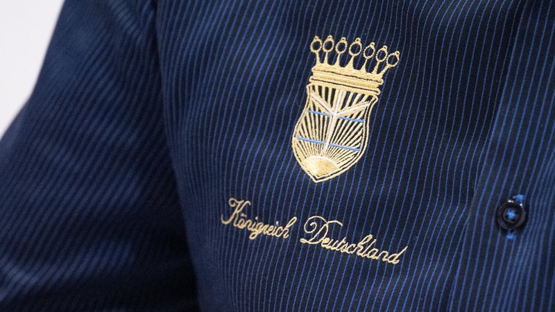 Ein schwarzes Hemd mit blauen Streifen in einer Nahaufnahme. Darauf ist ein goldenes Wappen gestickt und die Aufschrift "Königreich Deutschland" | Bild: dpa-Bildfunk/Hendrik Schmidt Ein schwarzes Hemd mit blauen Streifen in einer Nahaufnahme. Darauf ist ein goldenes Wappen gestickt und die Aufschrift "Königreich Deutschland"