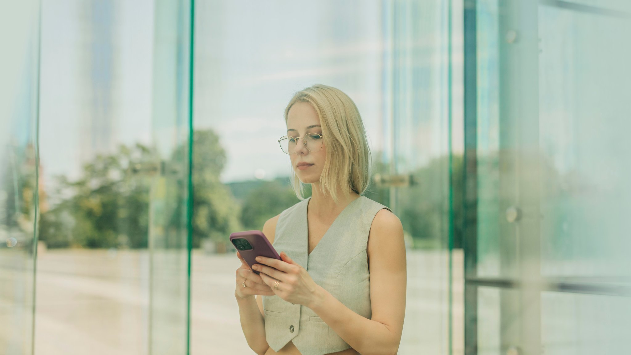 Blonde Frau mit Smartphone hinter Glas; Ständig erreichbar im Job: Das sind laut Wissenschaft die Folgen